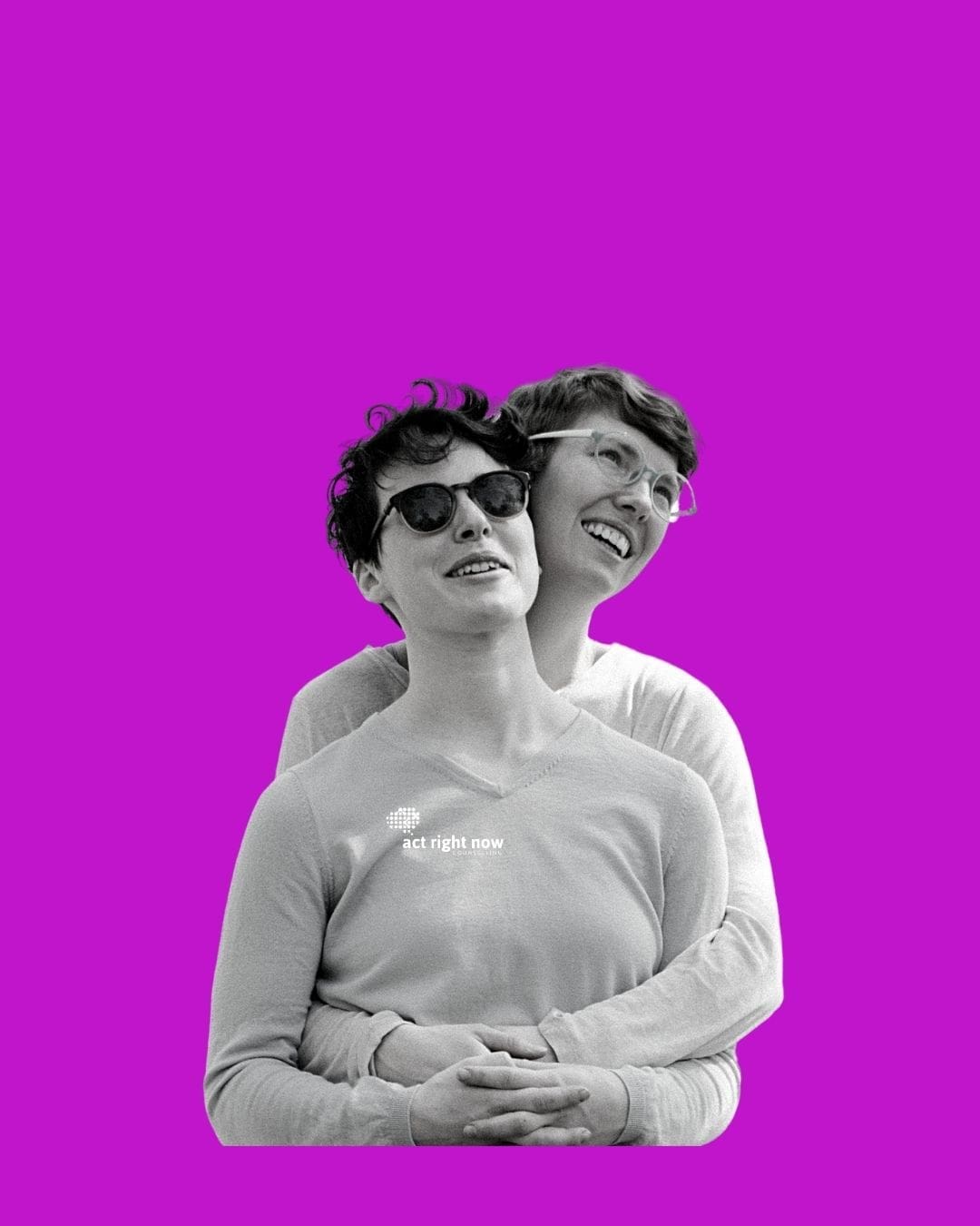 Smiling same-sex couple outdoors, one wearing sunglasses while being hugged from behind